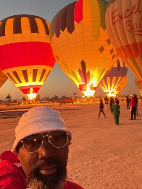 A man taking a selfie in front of a group of hot air balloons.